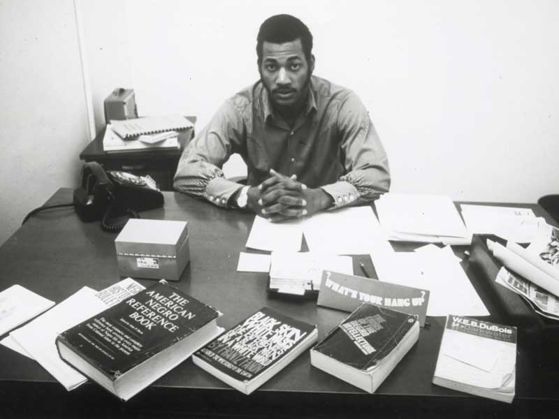 a student from the 1960s sitting behind a desk with books such as 'the american negro reference book' and a book by w.e.b. dubois