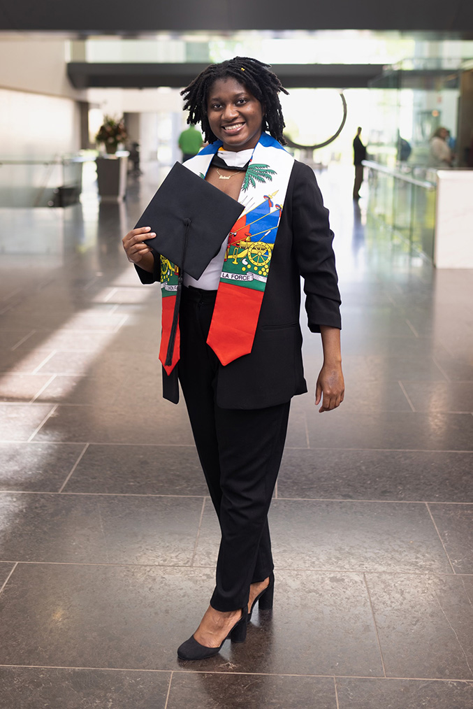 Dulanda Saintcyr stands in a lobby holding a graduation cap and wearing a stole