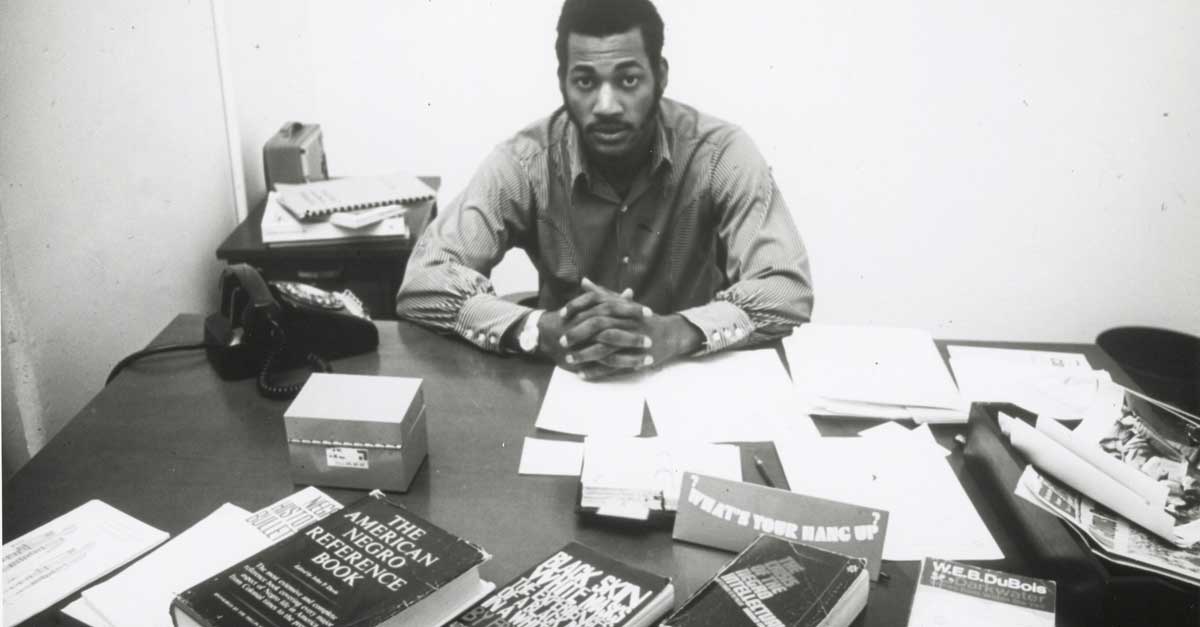 a student from the 1960s sitting behind a desk with books such as 'the american negro reference book' and a book by w.e.b. dubois